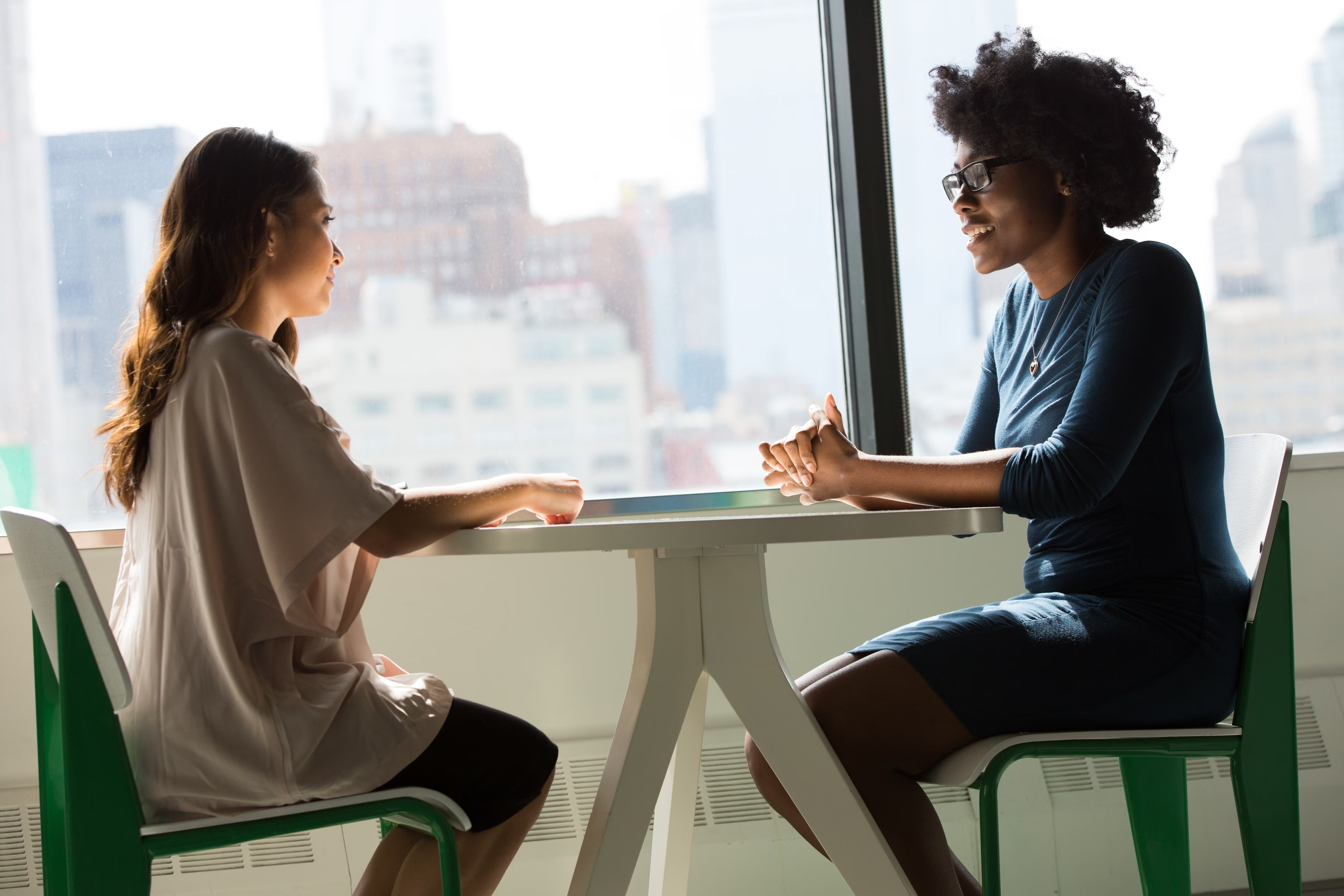 Two women sitting at a modern table talking with a city view behind them.